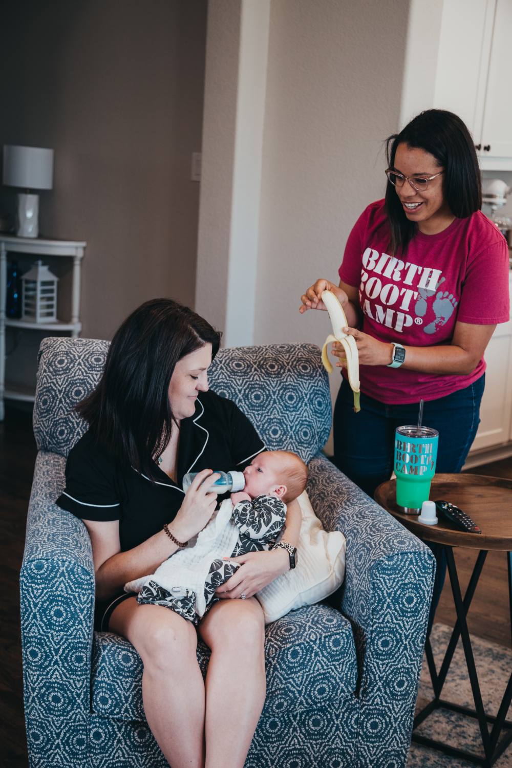 a new mom sitting in a chair feeding her newborn baby a bottle while her postpartum doula brings her a snack