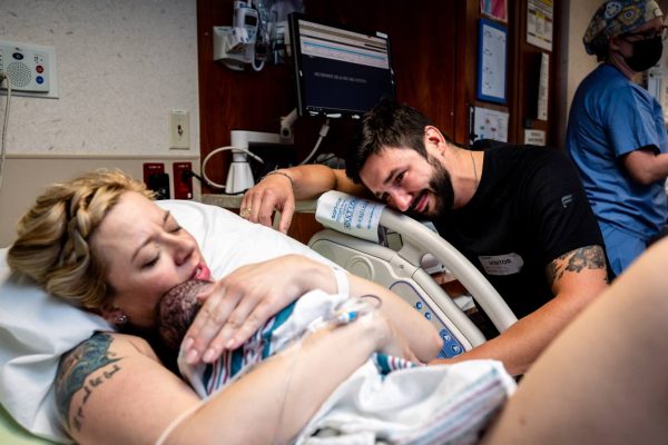 Woman lying in a hospital bed holding her newborn baby after giving birth while her husband looks admiringly at both of them