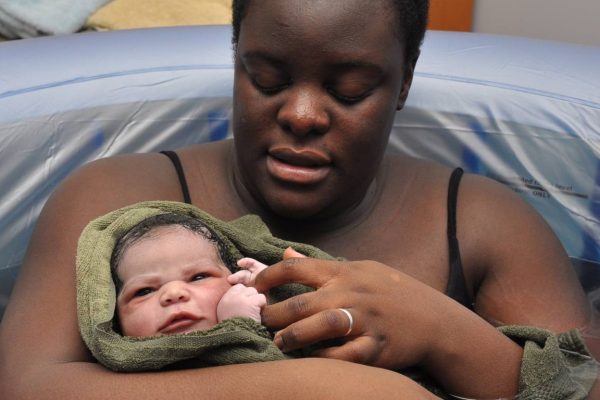 Black woman holding her wide eyed newborn baby in the birth tub immediately after having a natural water birth
