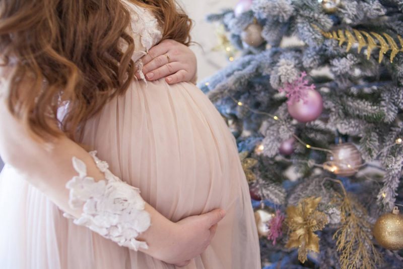 Pregnant woman wearing a lacy and flowy pink dress. All that is shown is her long curly red hair and her holding her beautiful baby bump. Flocked christmas tree is in the background decorated with pink and gold ornaments and decor. 