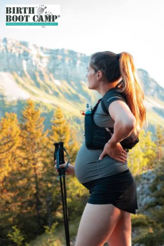 Pregnant woman overlooking a mountain scene. She is dressed in light clothing and is holding walking sticks in her right hand. 