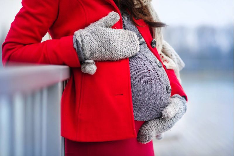 Pregnant women outside wearing a red winter coat, gray gloves, gray buttoned sweater ,and a gray scarf. She has one hand above her belly and one gloved hand below. 