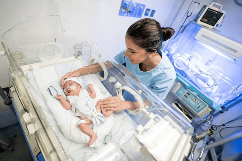 Nurse wearing blue scrubs is assisting a NICU baby in an incubator. 